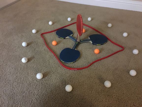 A ring of white ping pong balls surrounding 3 orange balls and 3 blue paddles in overlapping triangles. In the center, a red paddle facing upward.